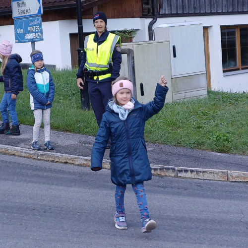 Ein Mädchen geht mit erhobener Hand über die Straße während die anderen Kinder und die Polizistin dabei zusehen