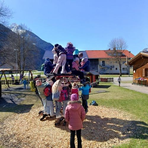 Viele Kinder auf einem Klettergerüst auf einem Spielplatz, im Hintergrund blauer Himmel und Häuser