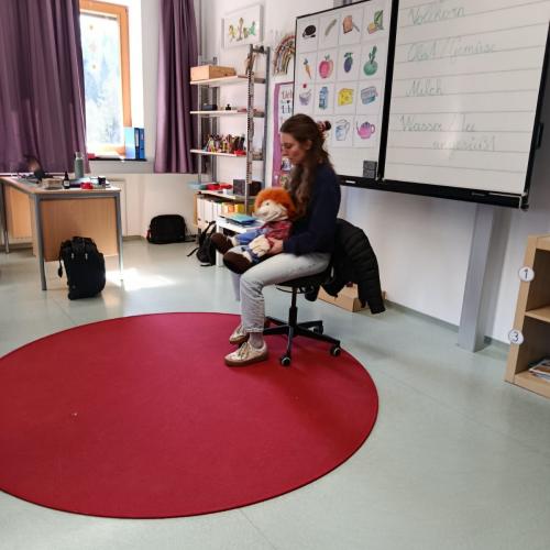 Vortragende mit Handpuppe auf Stuhl vor weißer Tafel