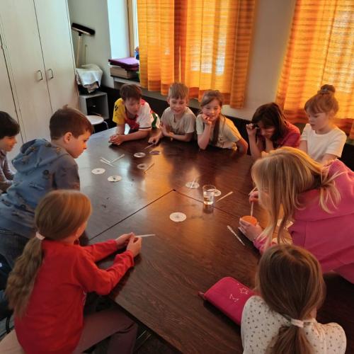 Eine blonde Frau hantiert mit einer Pipette an einem großen Tisch umgeben von einigen Kindern 