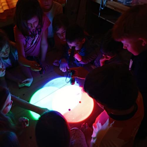 Gruppe von Schülern steht um Tisch, Frau leuchtet mit verschieden farbigen Taschenlampen auf den Tisch