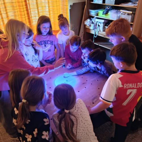 Gruppe von Schülern steht um Tisch, Frau leuchtet mit verschieden farbigen Taschenlampen auf den Tisch