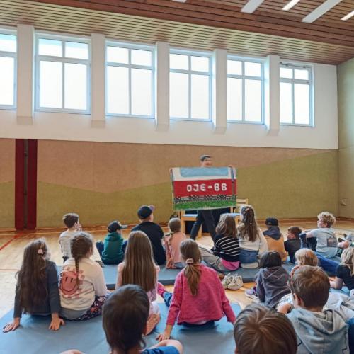 Kinder am Boden sitzend im Turnsaal, Kabarettistin hält ein Schild
