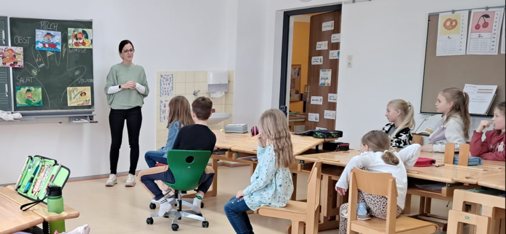 Workshop-Leiterin steht vor Tafel, Schülerinnen und Schüler sitzen auf ihren Plätzen und schauen und hören zu