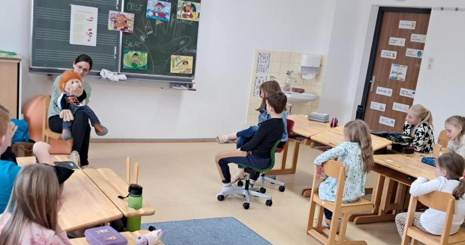 Klassenzimmer, Workshop-Leiterin sitzt mit großer Puppe vor Tafel, Kinder schauen und hören zu auf ihren Plätzen