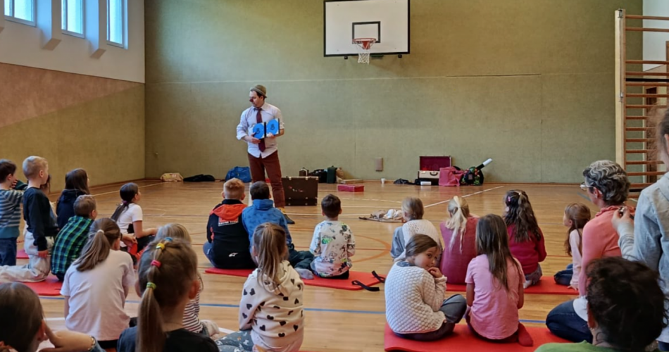 Ein Clown steht im Turnsaal vor sitzenden Kindern