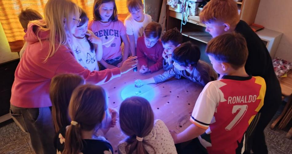 Gruppe von Schülern steht um Tisch, Frau leuchtet mit verschieden farbigen Taschenlampen auf den Tisch