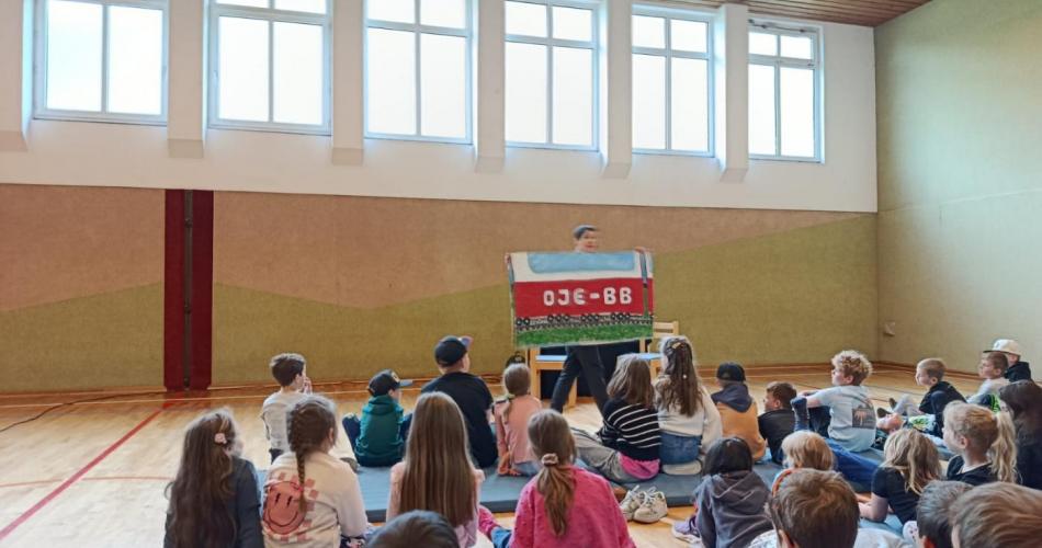 Kinder am Boden sitzend im Turnsaal, Kabarettistin hält ein Schild